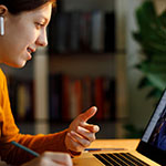 a student seated at a table engages in a video conference on her laptop